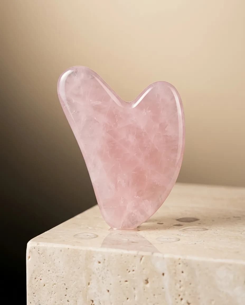 Rozenkwarts gua sha-massagesteen op een marmeren voetstuk in zacht studiolicht